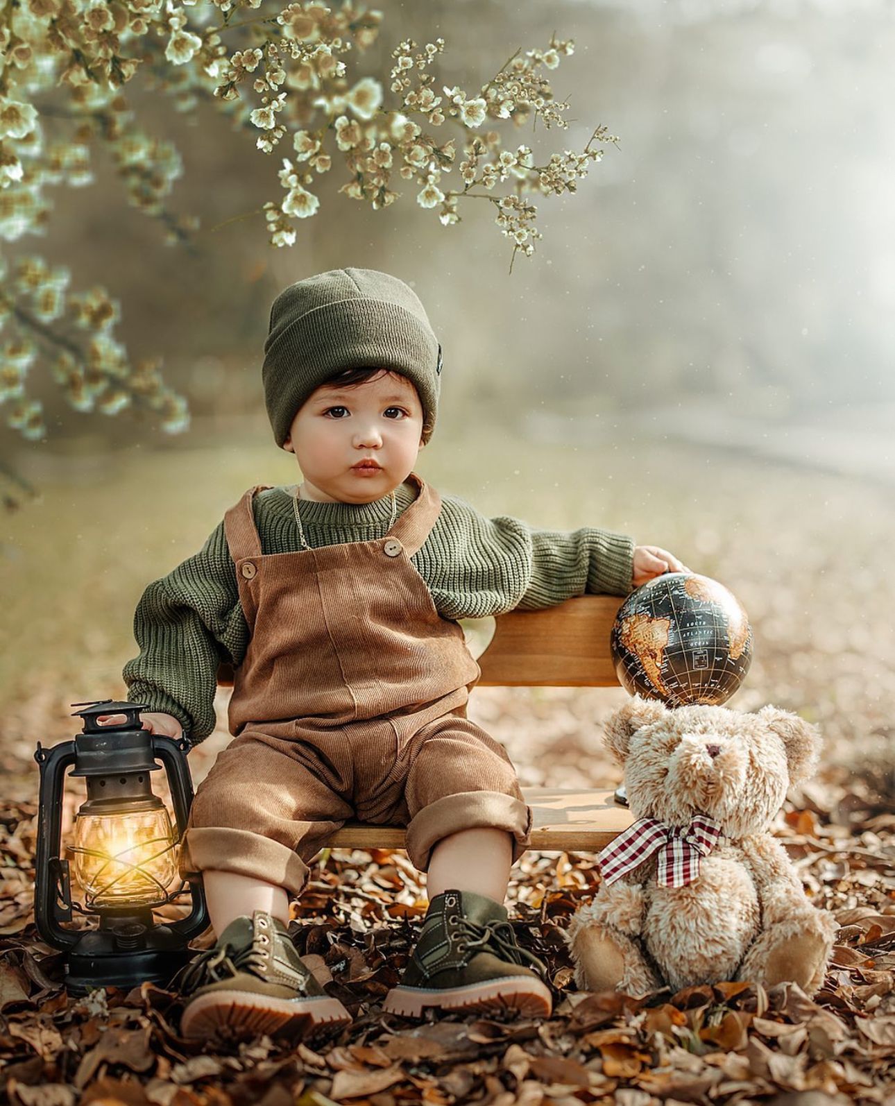 A toddler boy in brown overalls sits on autumn leaves beside a glowing lantern and teddy bear under flowering branches.