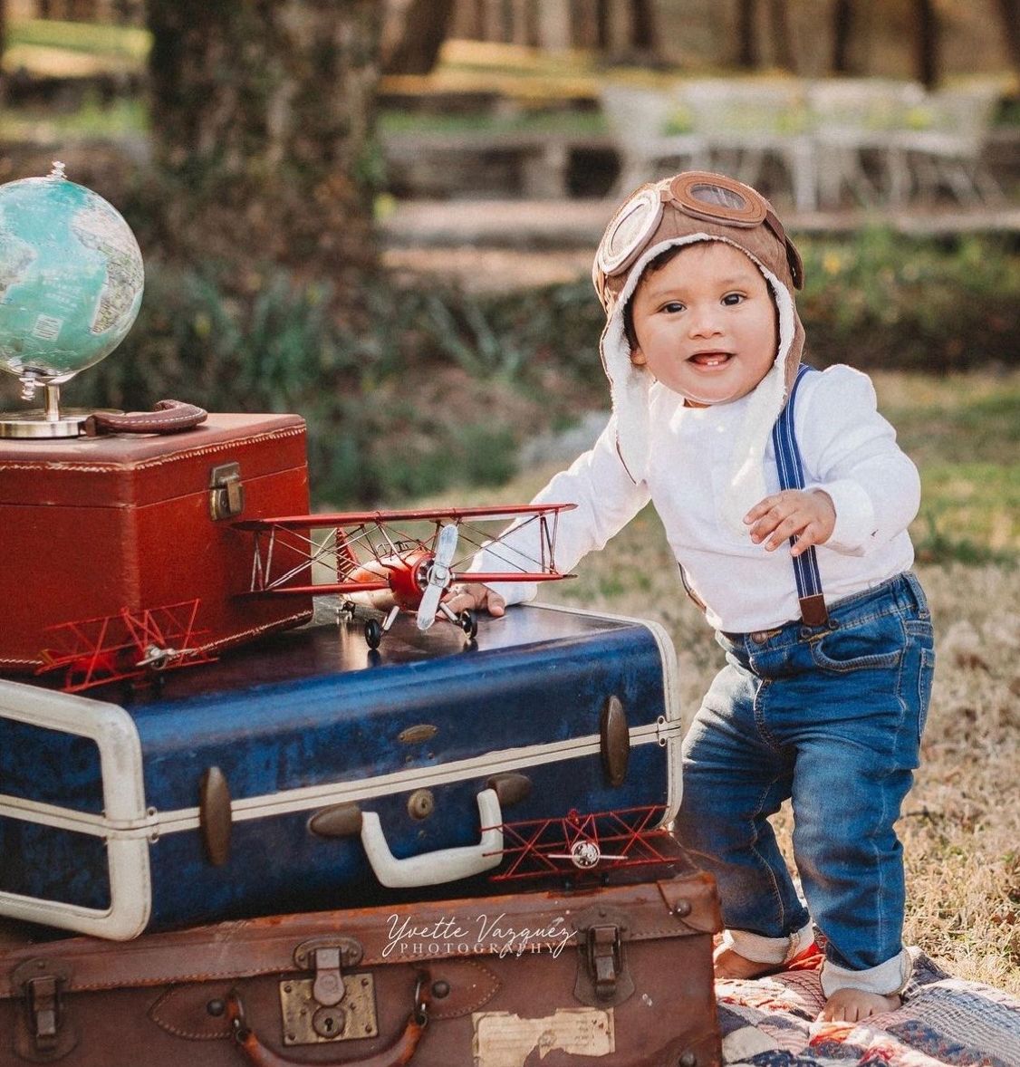 Toddler in aviator hat and goggles stands by vintage suitcases with globe and toy biplane outdoors.