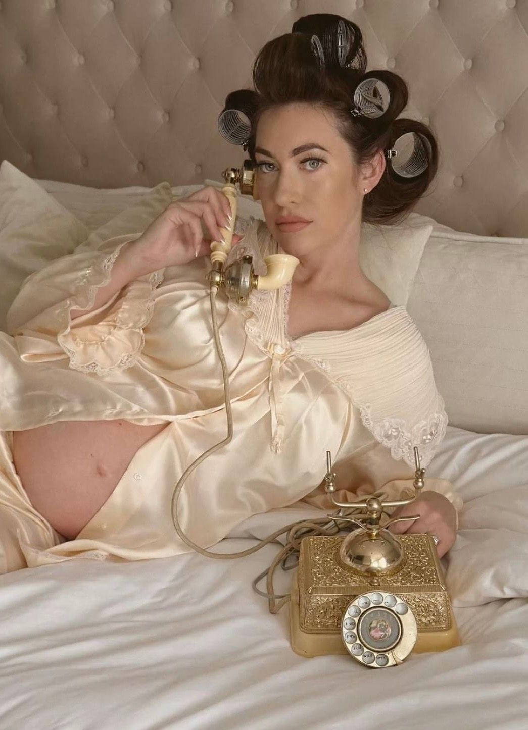 Pregnant woman in satin robe with hair rollers talking on vintage rotary phone while reclining on bed.