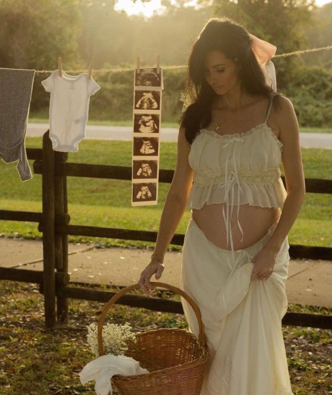 Pregnant woman in white dress holding basket outdoors near clothesline with baby clothes and ultrasound photos.
