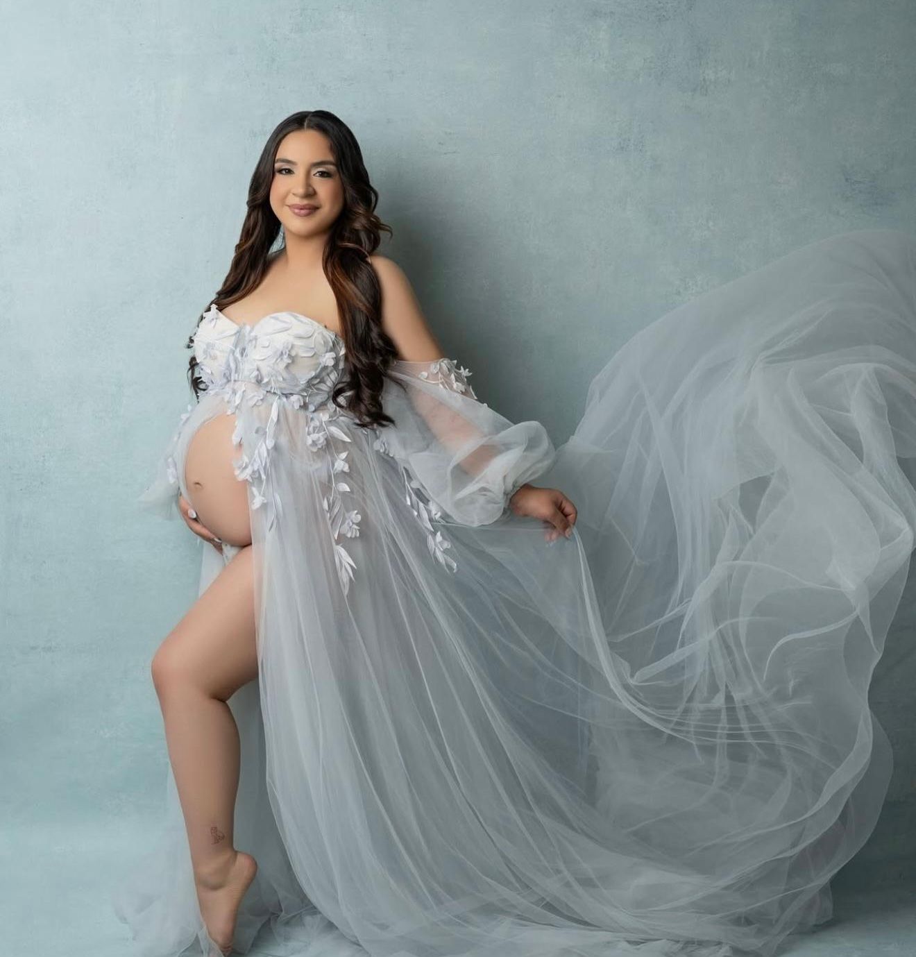 Pregnant woman in elegant white floral tulle maternity gown with flowing fabric, posing for professional maternity photo shoot.