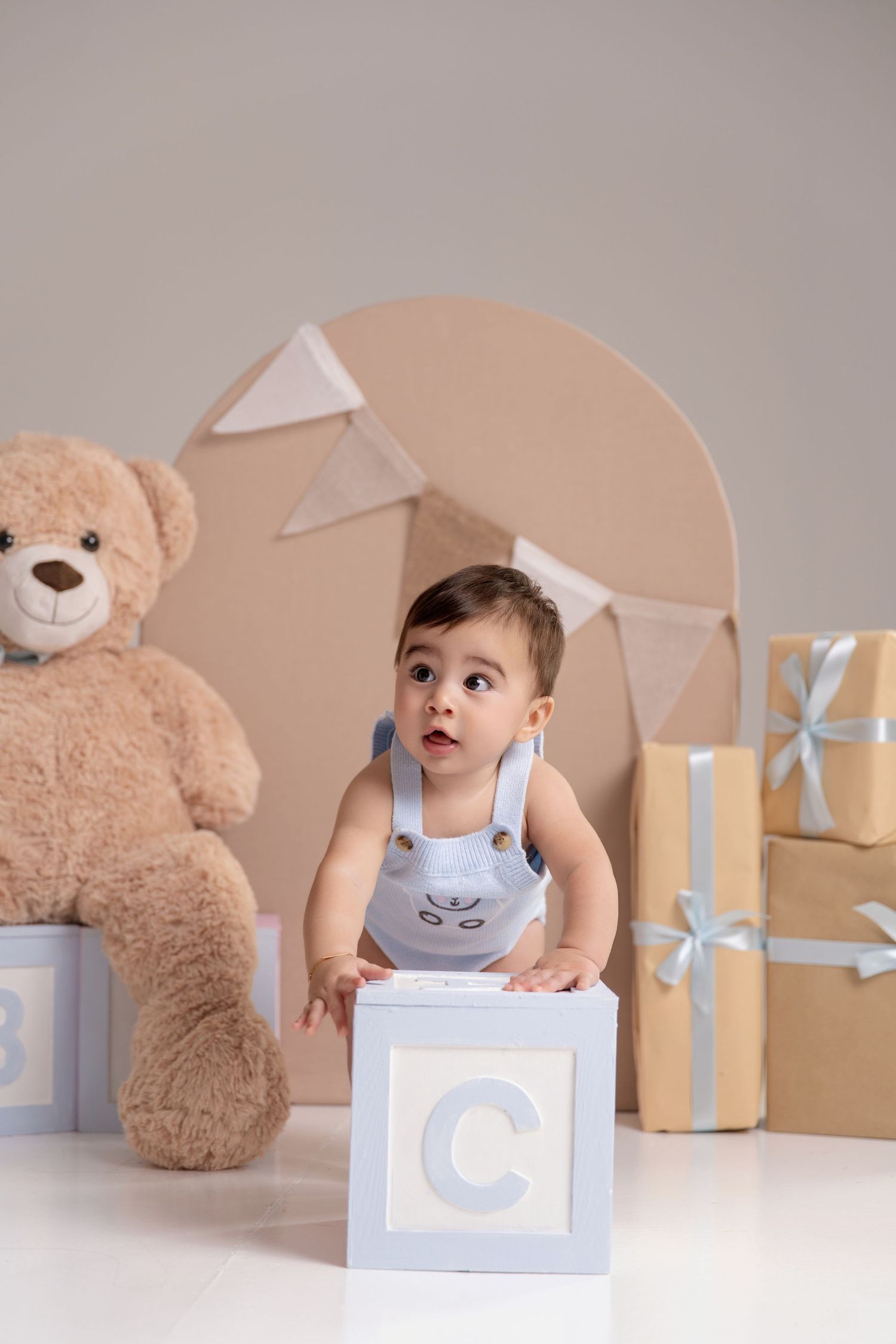 Cute baby boy in blue overalls leaning on alphabet block C, with teddy bear and gifts in background.