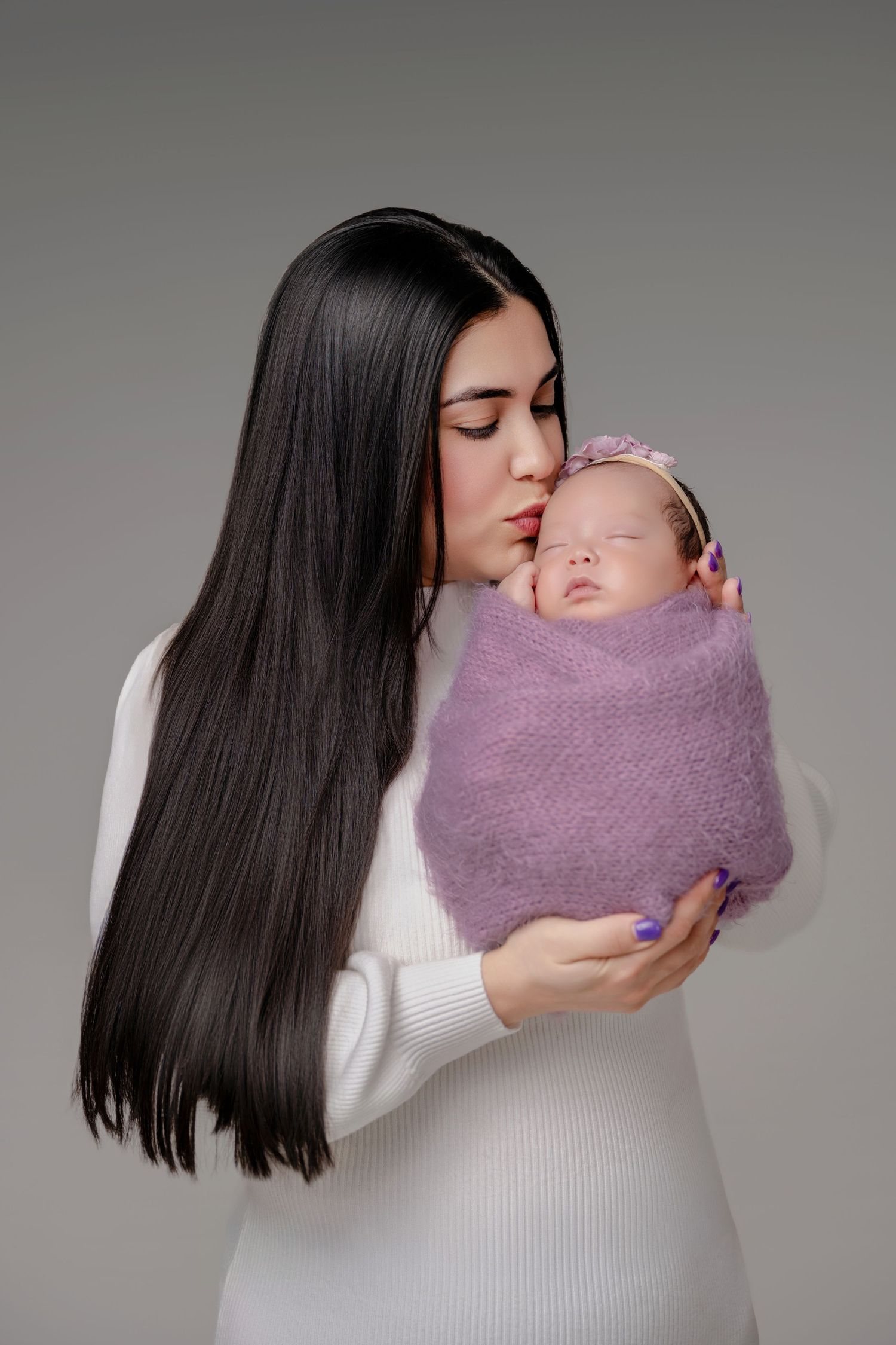 A mother with long dark hair in a white dress gently kisses her sleeping newborn wrapped in a purple blanket.