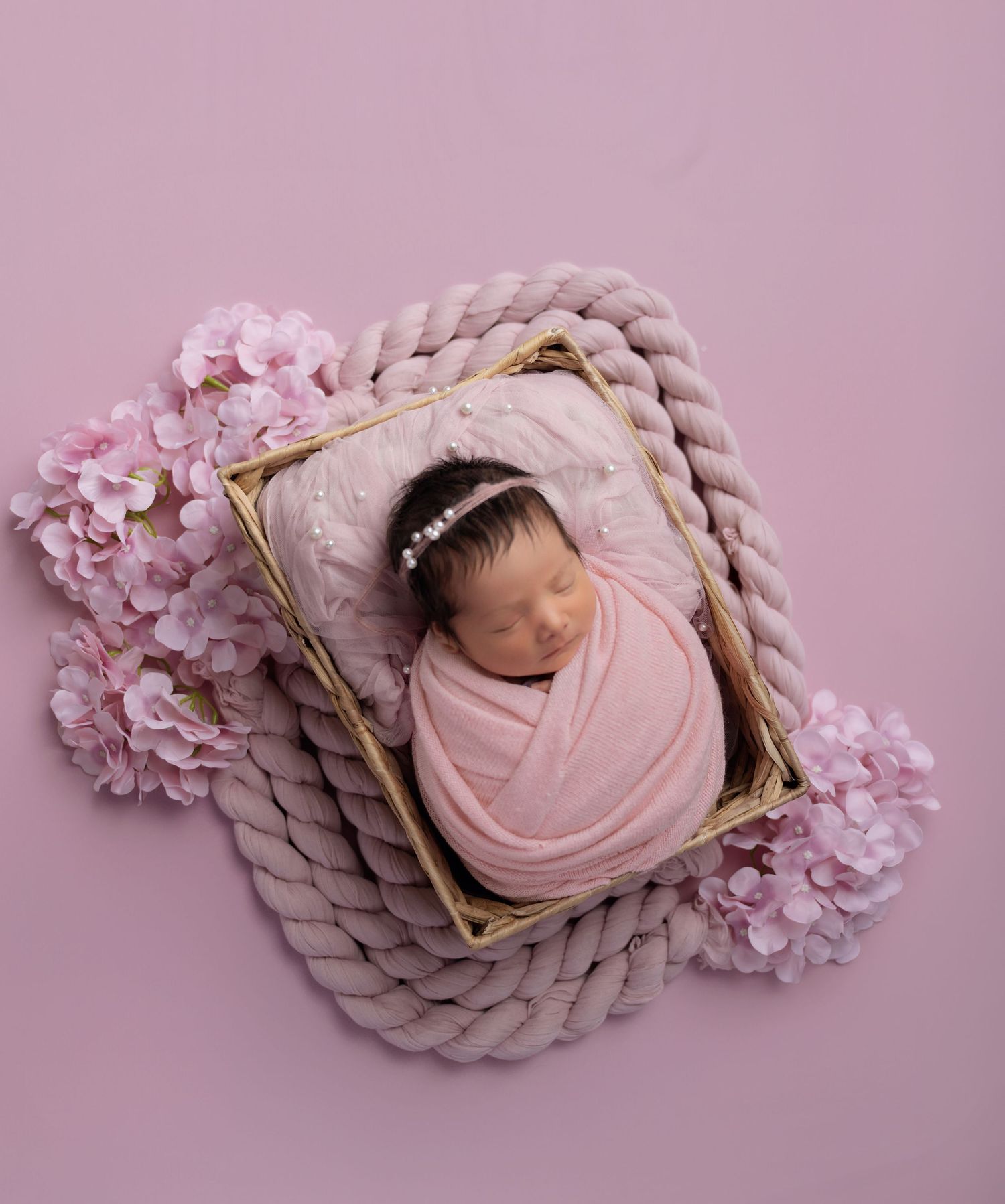 Newborn baby girl swaddled in pink wrap, sleeping in a golden frame basket with floral decorations on pink background.