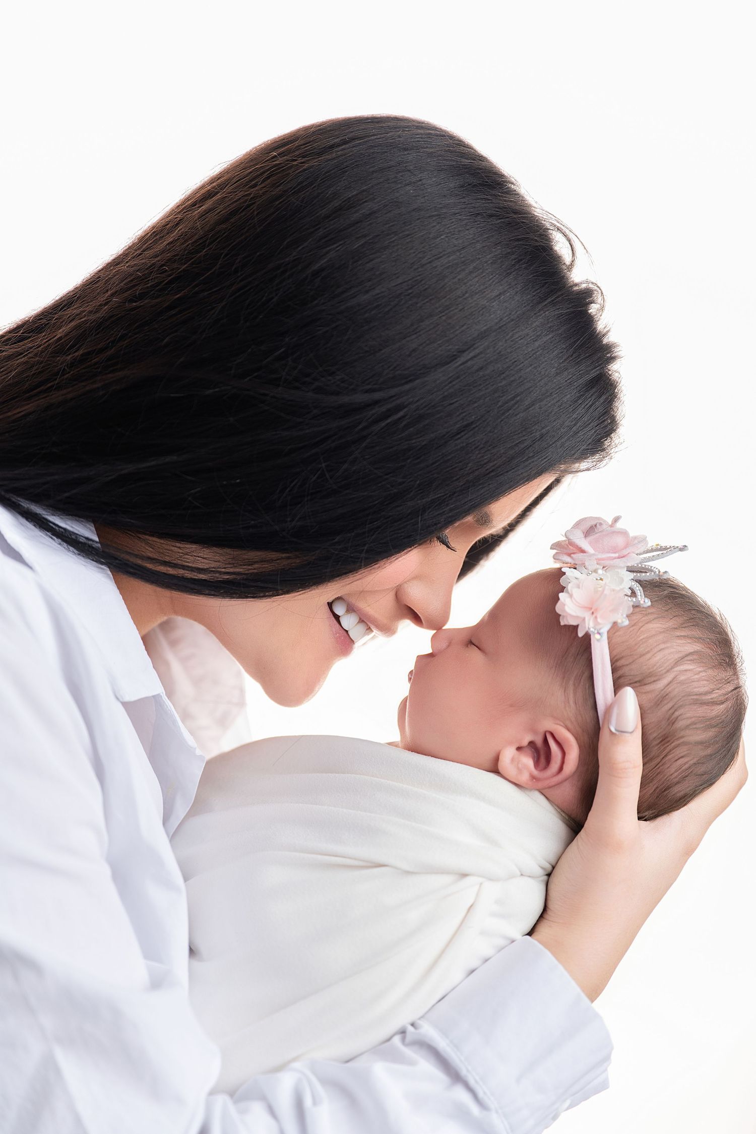 A smiling dark-haired mother in white gently holds her swaddled newborn baby girl with a floral headband, touching foreheads.
