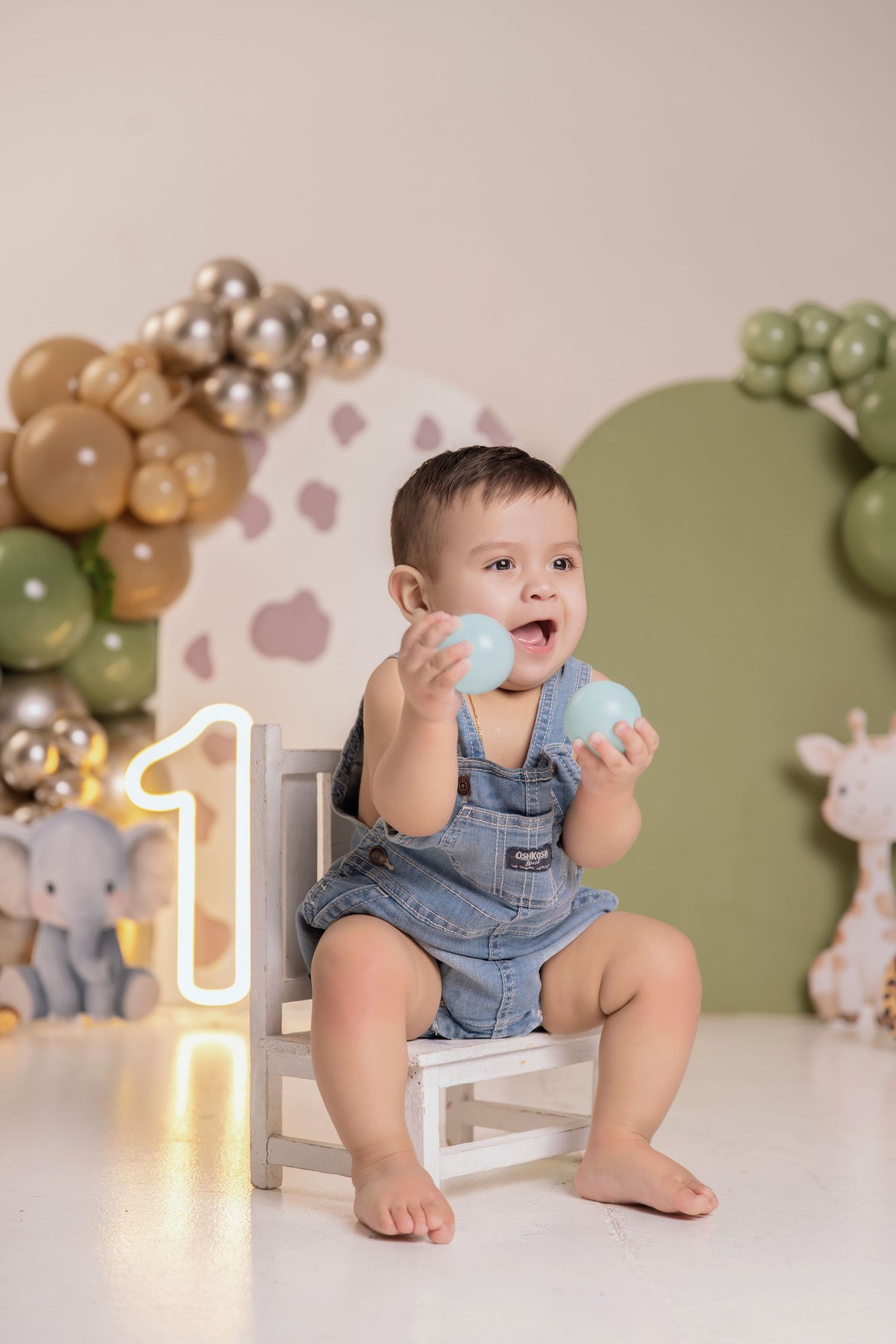 Baby boy in denim overalls sits on white chair holding blue balls at first birthday party with balloon garland backdrop.