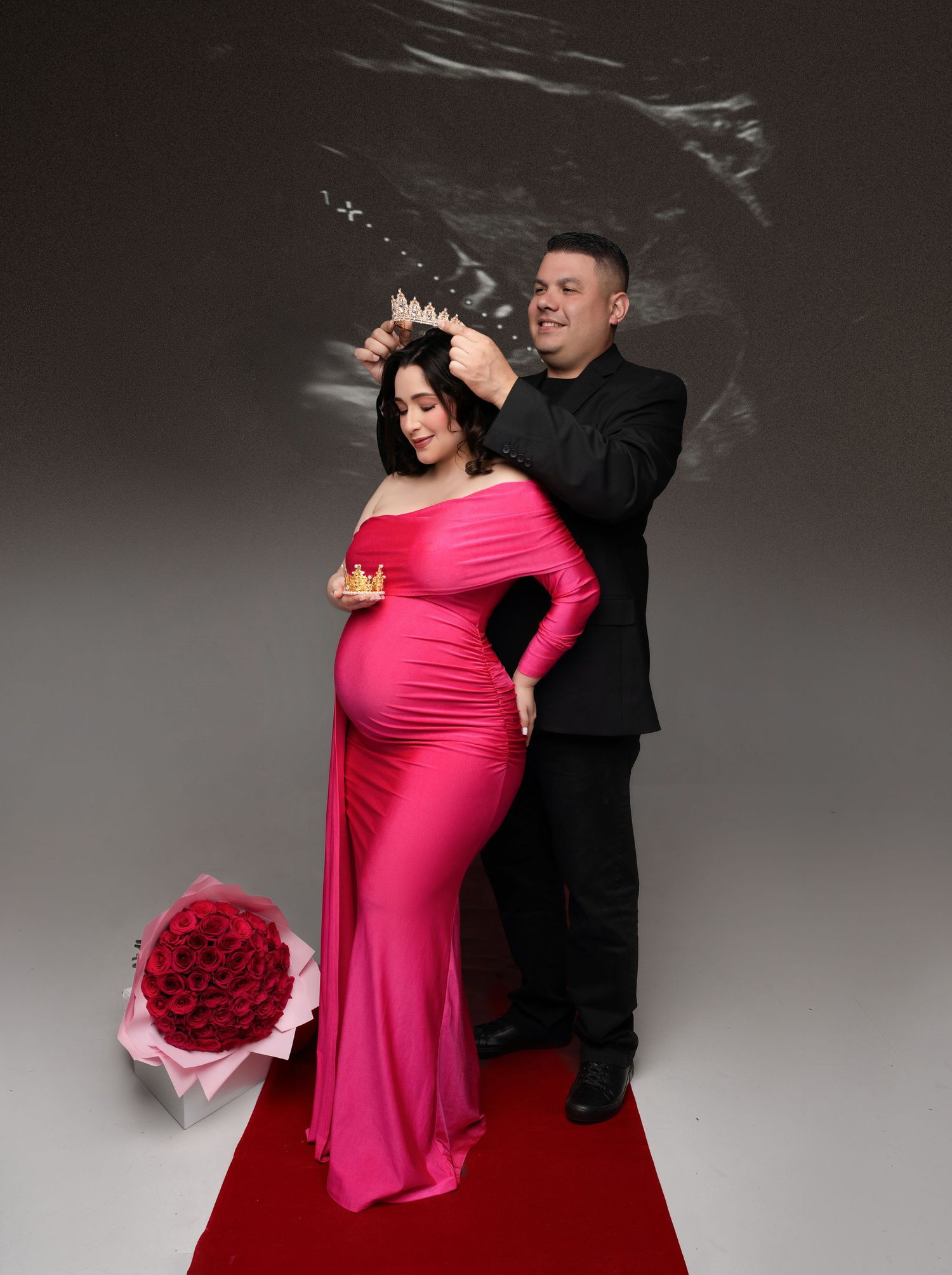A couple in formal attire performs an elegant dance pose against a dark studio backdrop with flowing pink fabric.