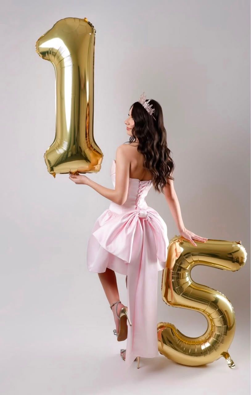 A young woman in a pink strapless dress and tiara poses with gold number 15 balloons for a quinceañera celebration.