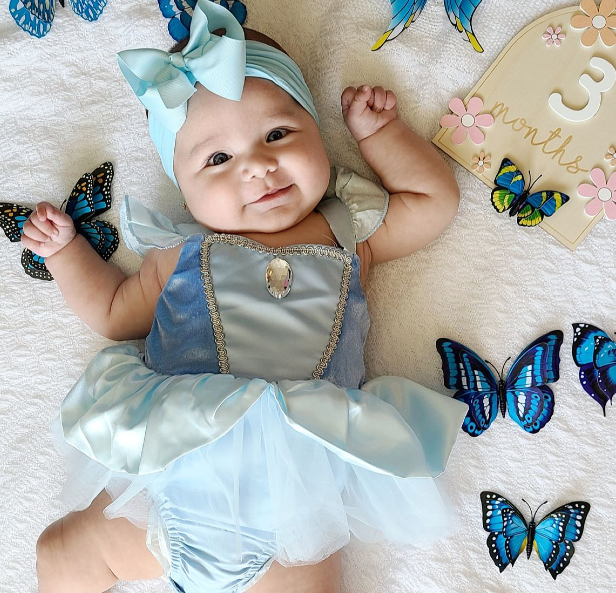 Adorable baby girl in Cinderella dress with blue bow headband surrounded by colorful butterflies at 3 months old.