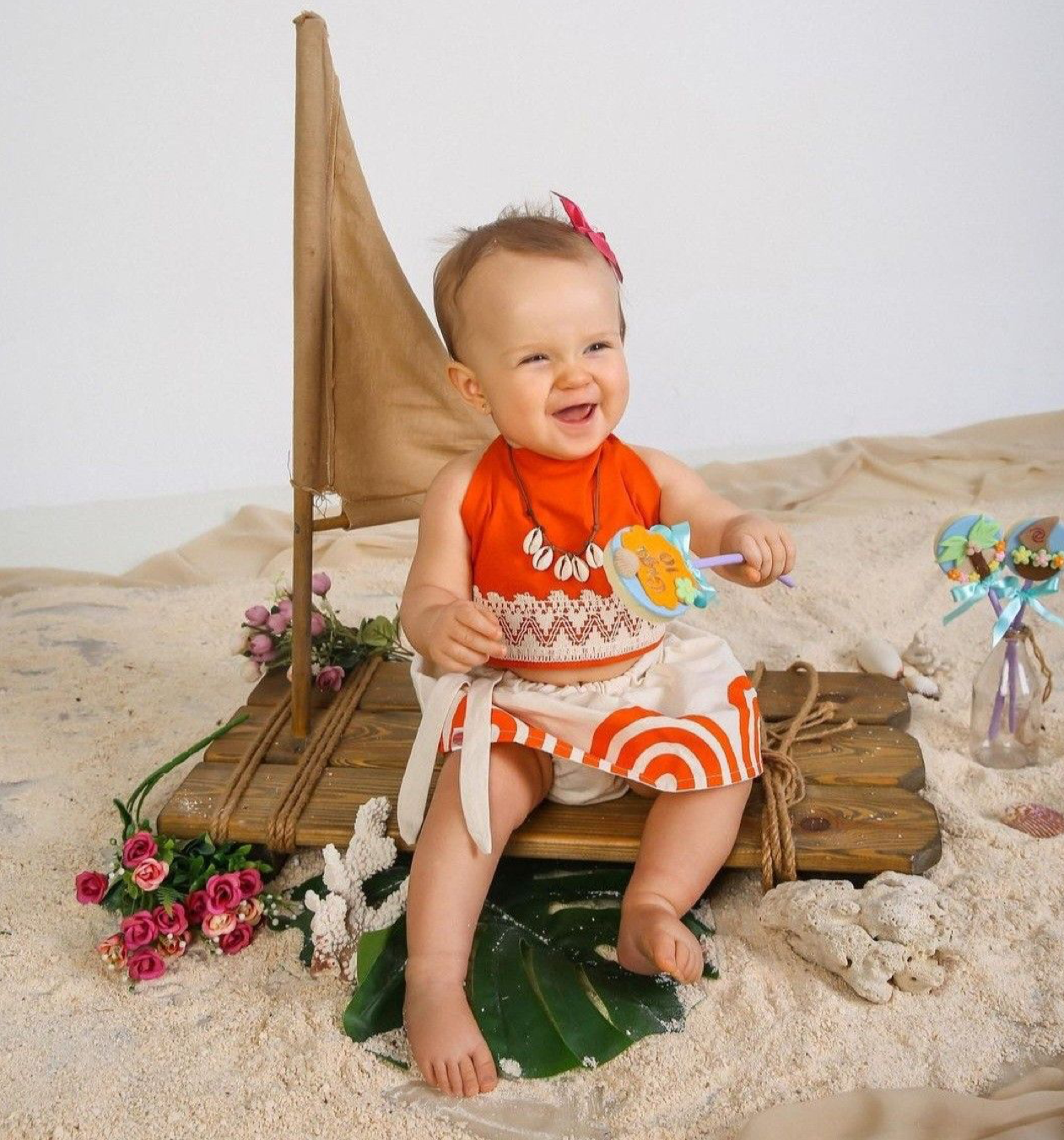 Baby girl in Moana costume sits smiling on sandy beach set with toy sailboat and tropical flowers.