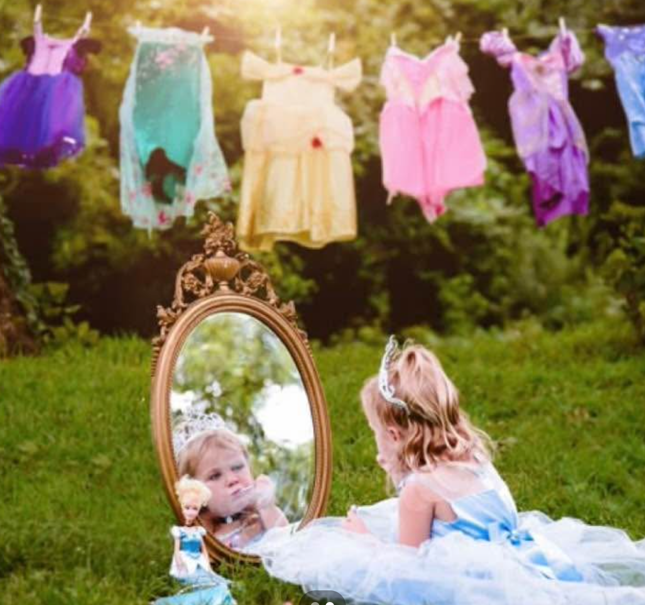 A little girl in a princess dress sits before a vintage mirror outdoors, with colorful dresses hanging on a line behind her.