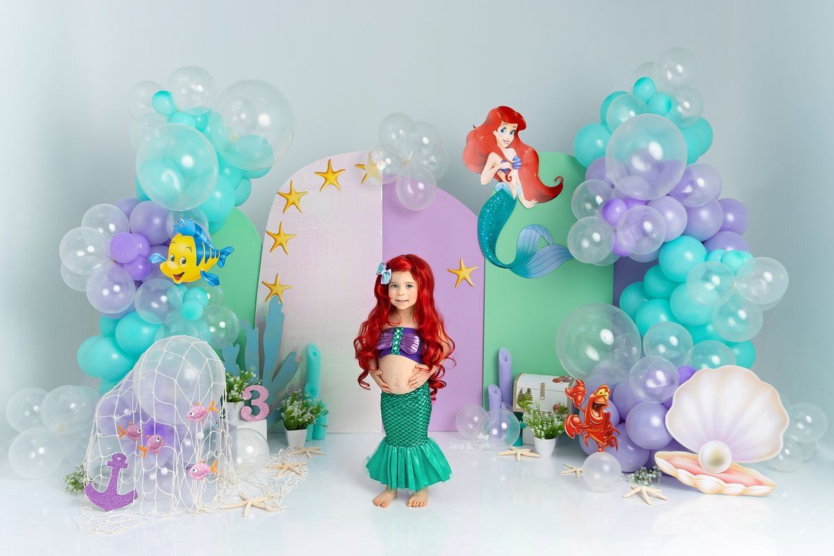 A little girl dressed as Ariel from The Little Mermaid poses with balloon arch and themed decorations.