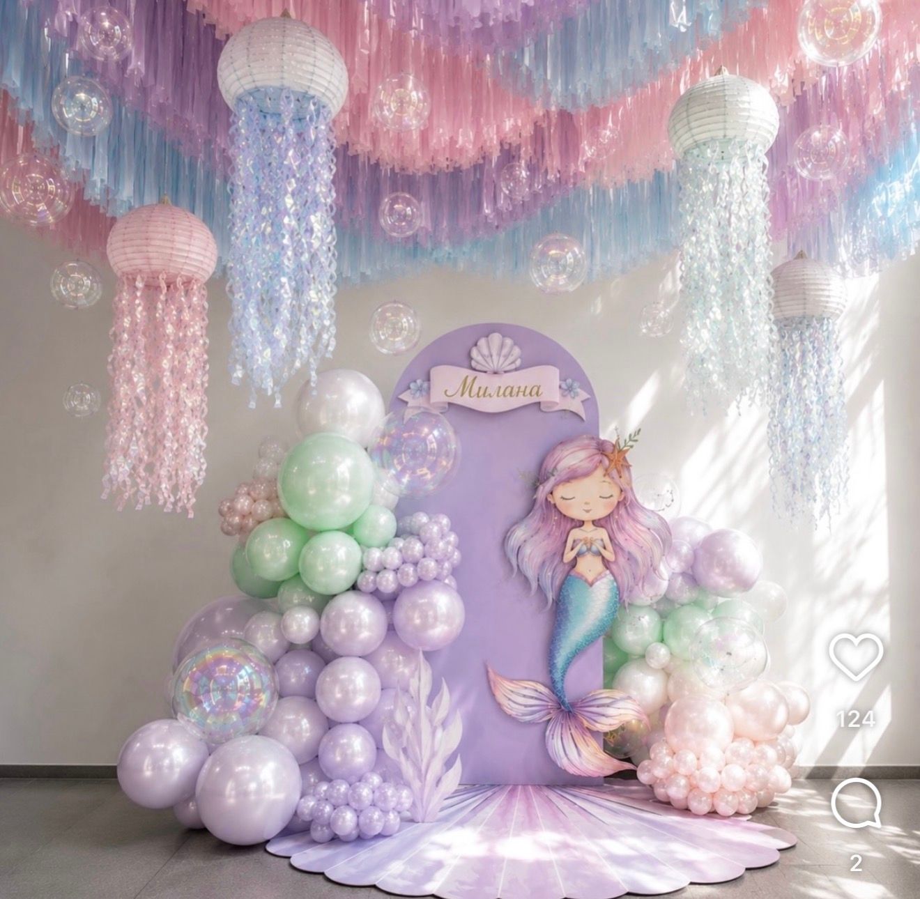 Mermaid themed birthday party decor with pastel balloons, jellyfish ceiling decorations, and a mermaid cutout backdrop.
