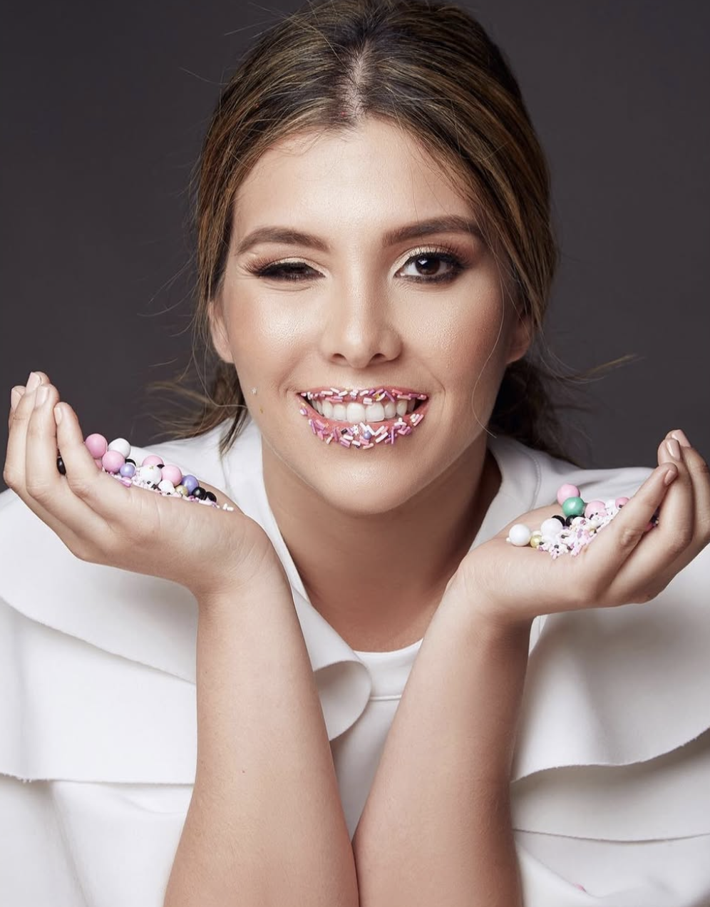 Young woman in white outfit smiling with colorful candy sprinkles on lips, holding pastel beads in both hands.