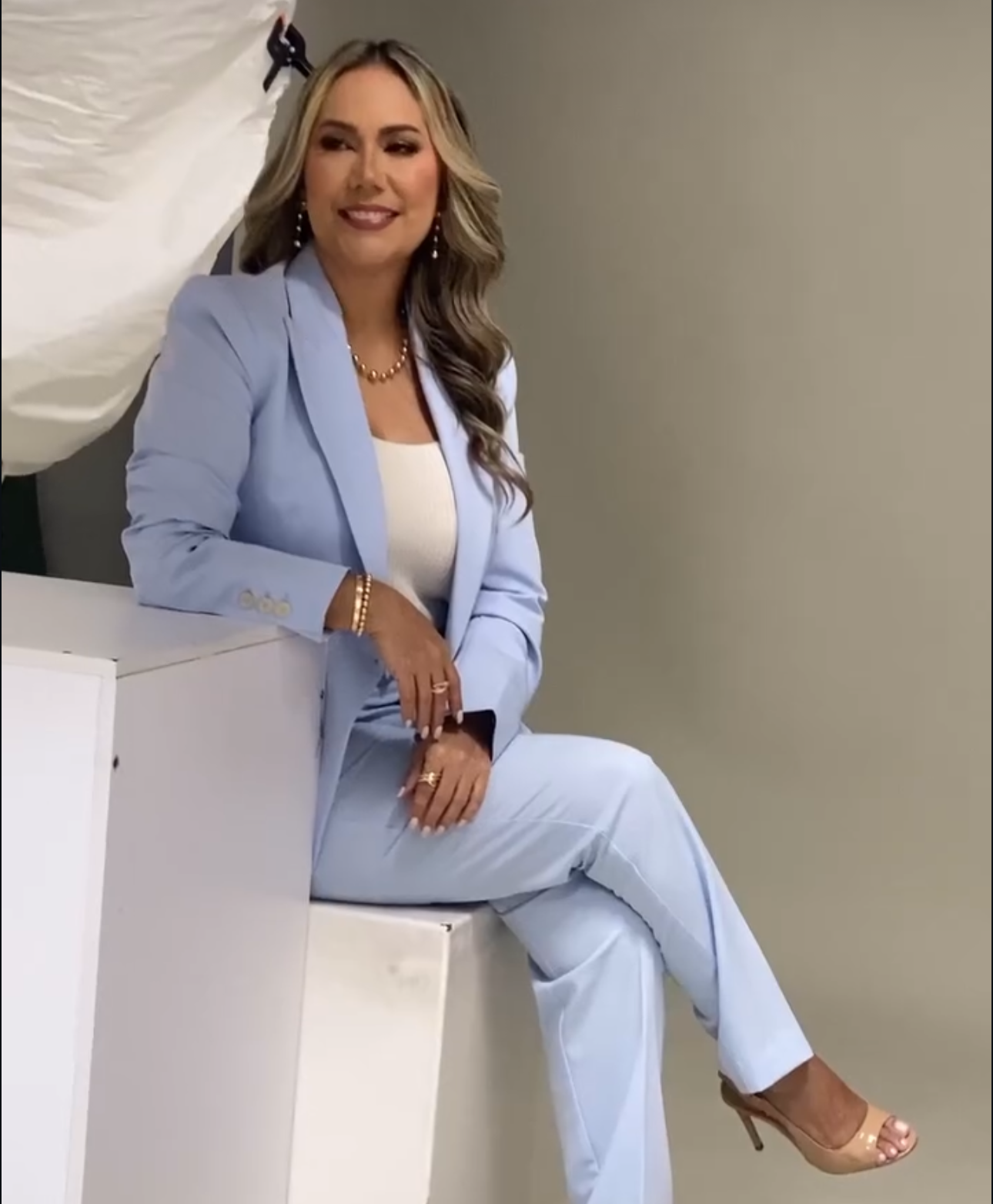 Woman in light blue pantsuit sitting confidently on white surface, wearing heels and gold jewelry.