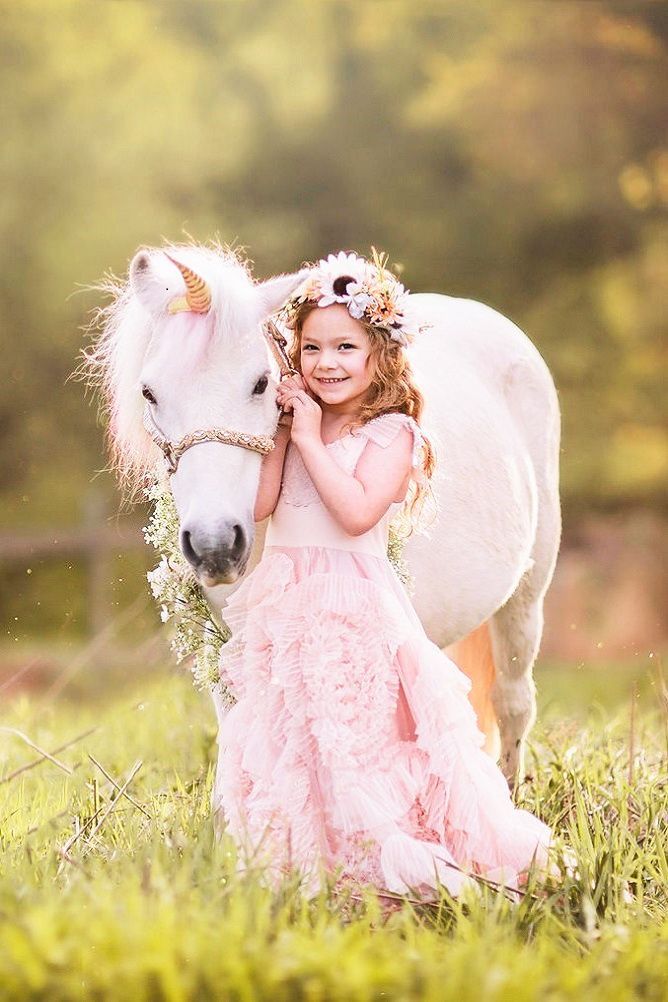 A young girl in a pink ruffled dress and flower crown smiles beside a white unicorn pony in a sunny meadow.