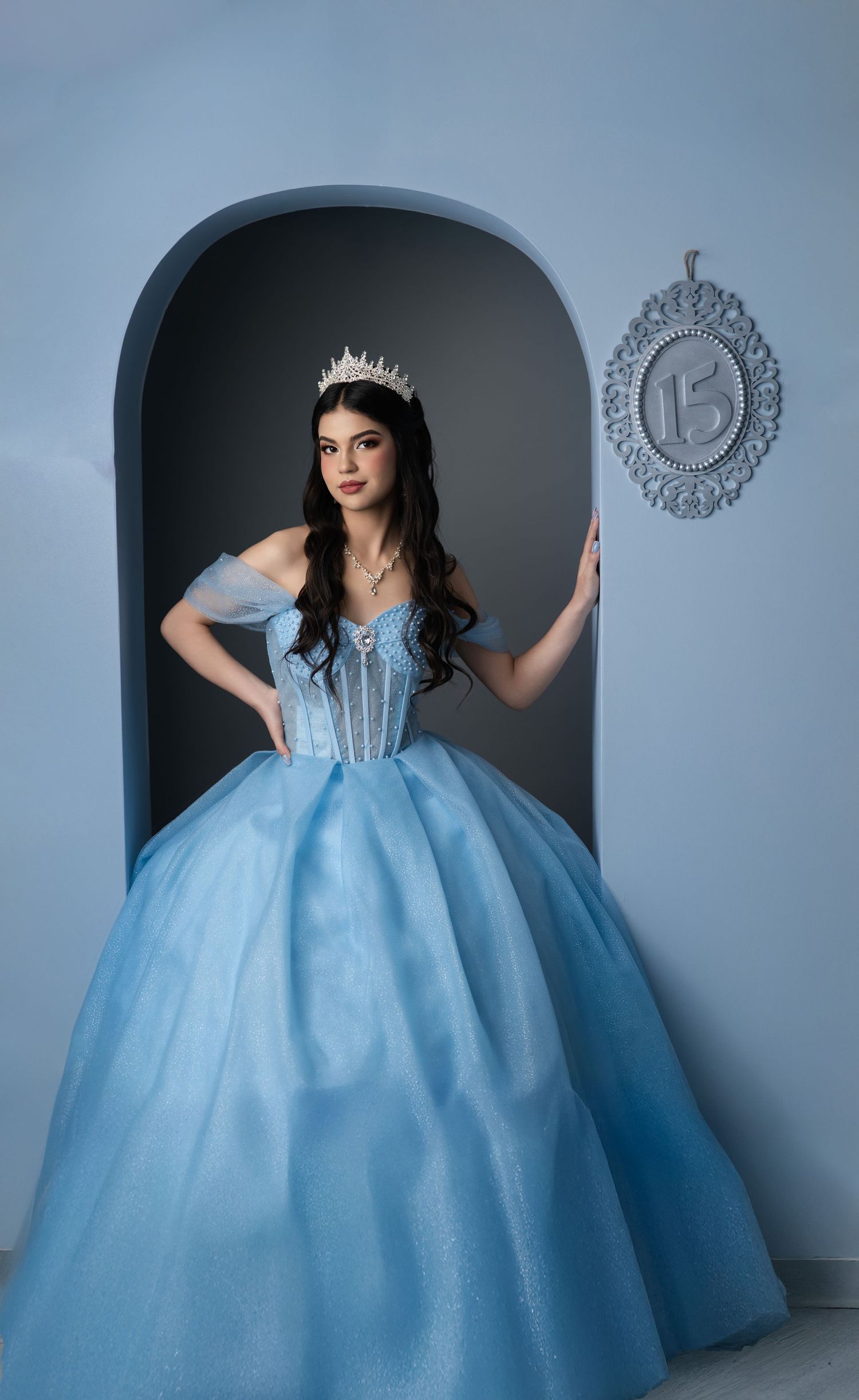 Young woman in a light blue ball gown and tiara poses elegantly beside a decorative number 15 sign for her Quinceañera.
