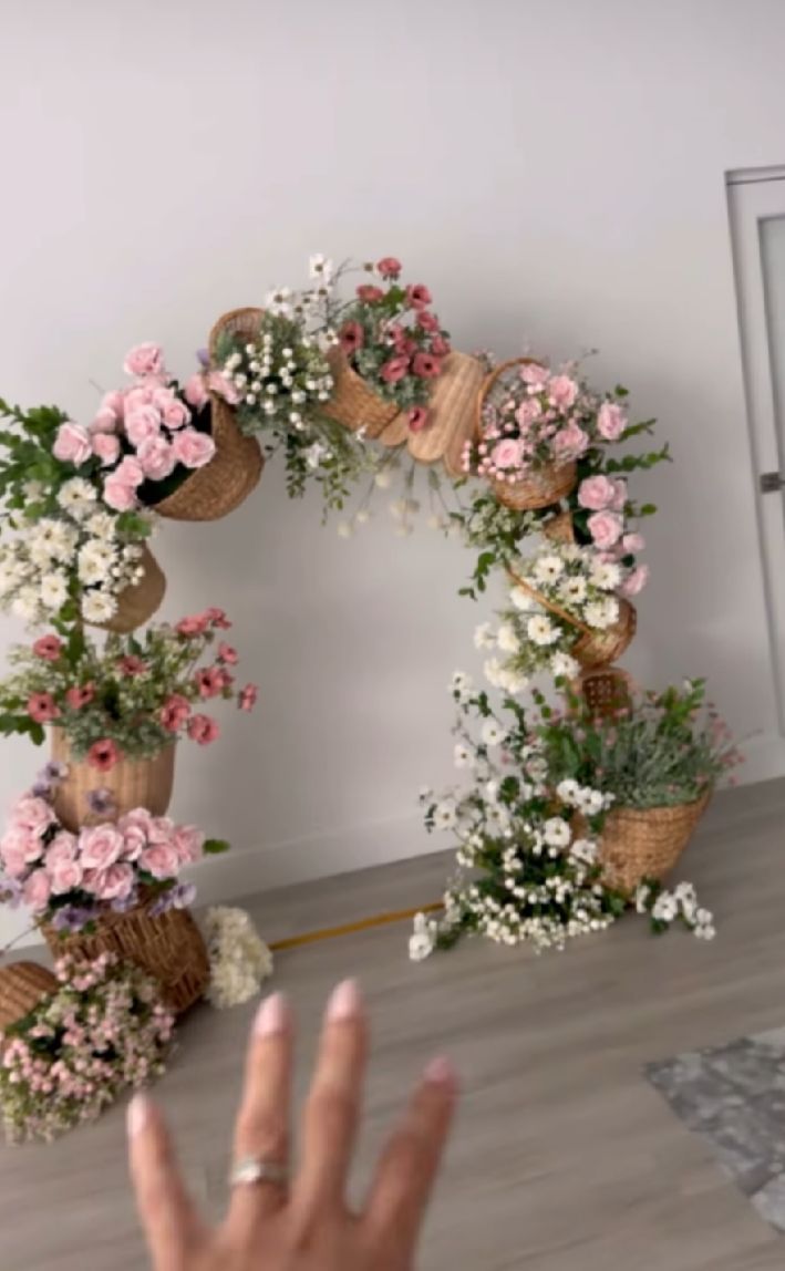 A hand gestures in front of a romantic floral arch made with pink roses, white baby's breath, and woven baskets.