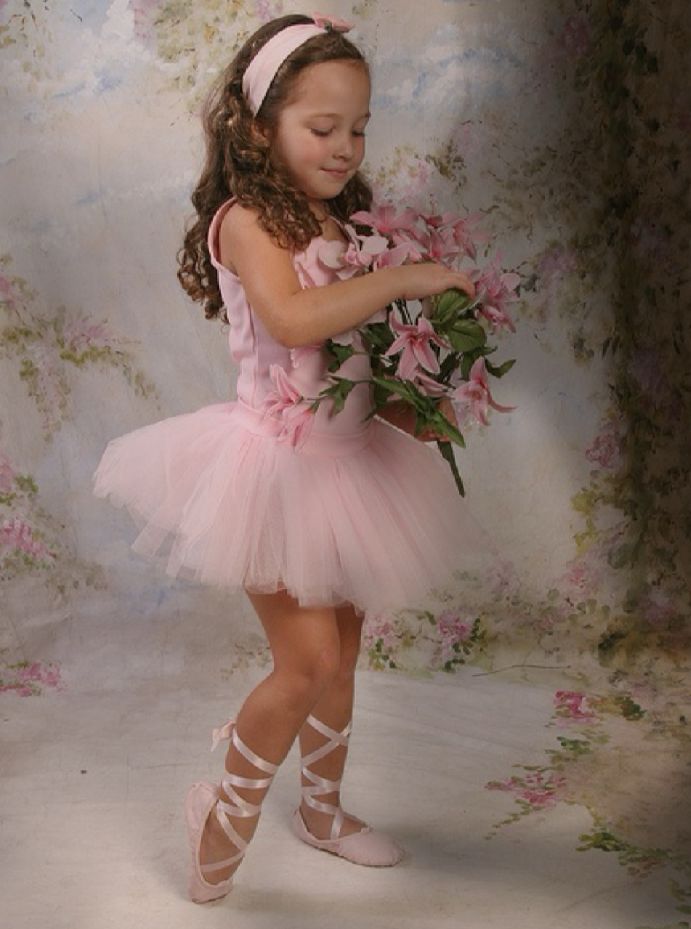 A young dancer in a pink tutu and ballet slippers poses gracefully with pink flowers against a vintage backdrop.