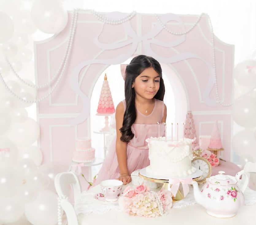 A dreamy tea party scene with pink decor features a white cake, floral arrangements and delicate teaware on display.