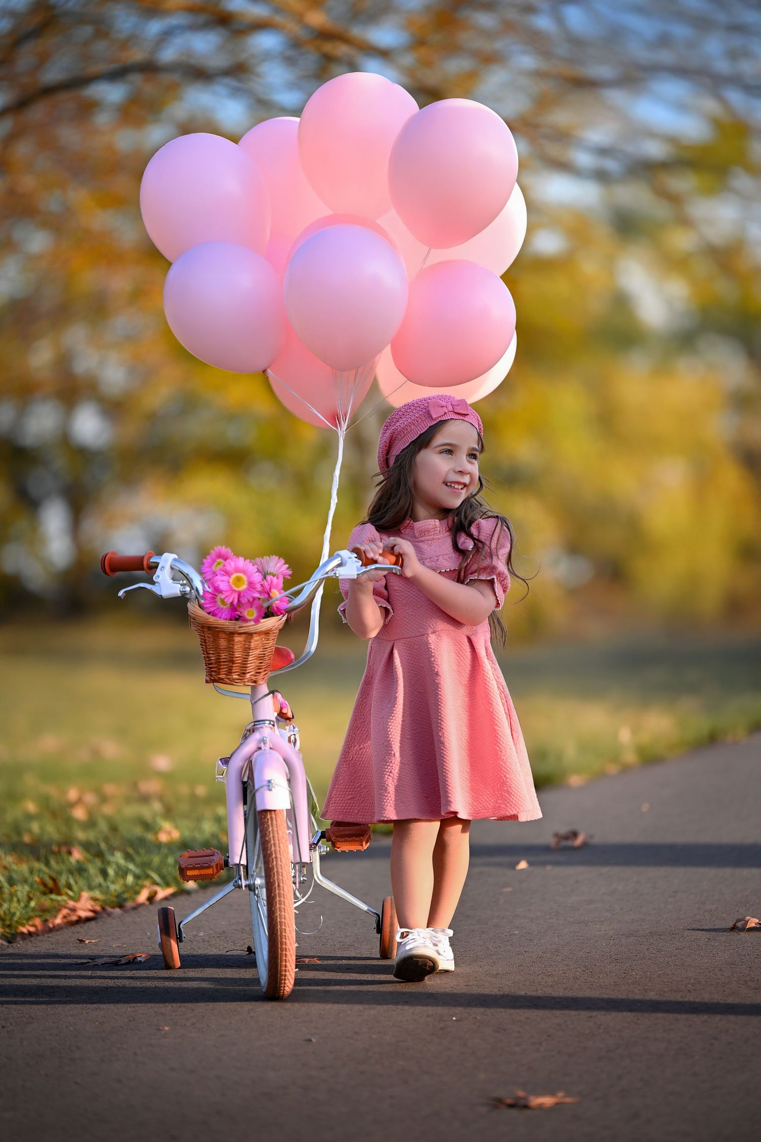 A little girl in a pink dress and beret stands on a path holding pink balloons beside a white bicycle with flowers.