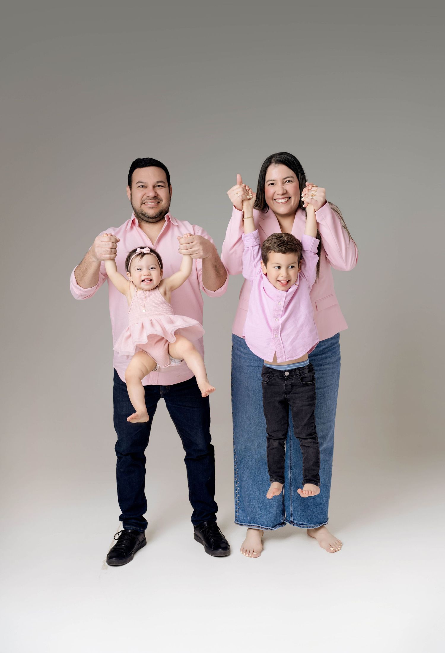 Family of four in matching pink shirts giving thumbs up, parents holding young children in a studio portrait.