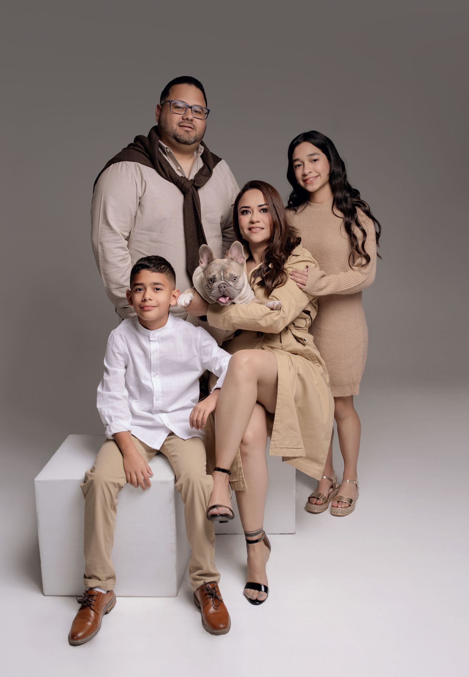Family of four posing with a French Bulldog in a professional studio portrait, wearing coordinated neutral tones.