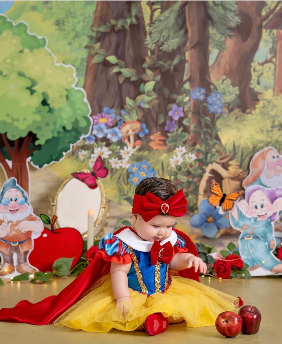 Baby girl dressed as Snow White in yellow and blue costume with red bow, posed with apples and Seven Dwarfs backdrop.