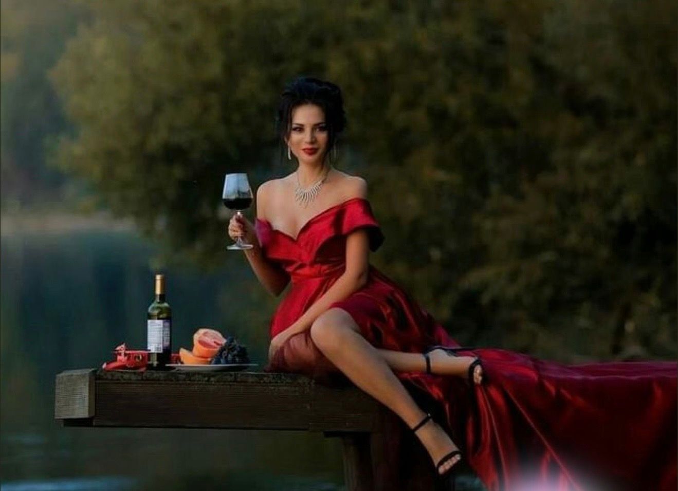 Woman in red off-shoulder gown holding wine glass on dock, with bottle and fruit, lush green trees background.