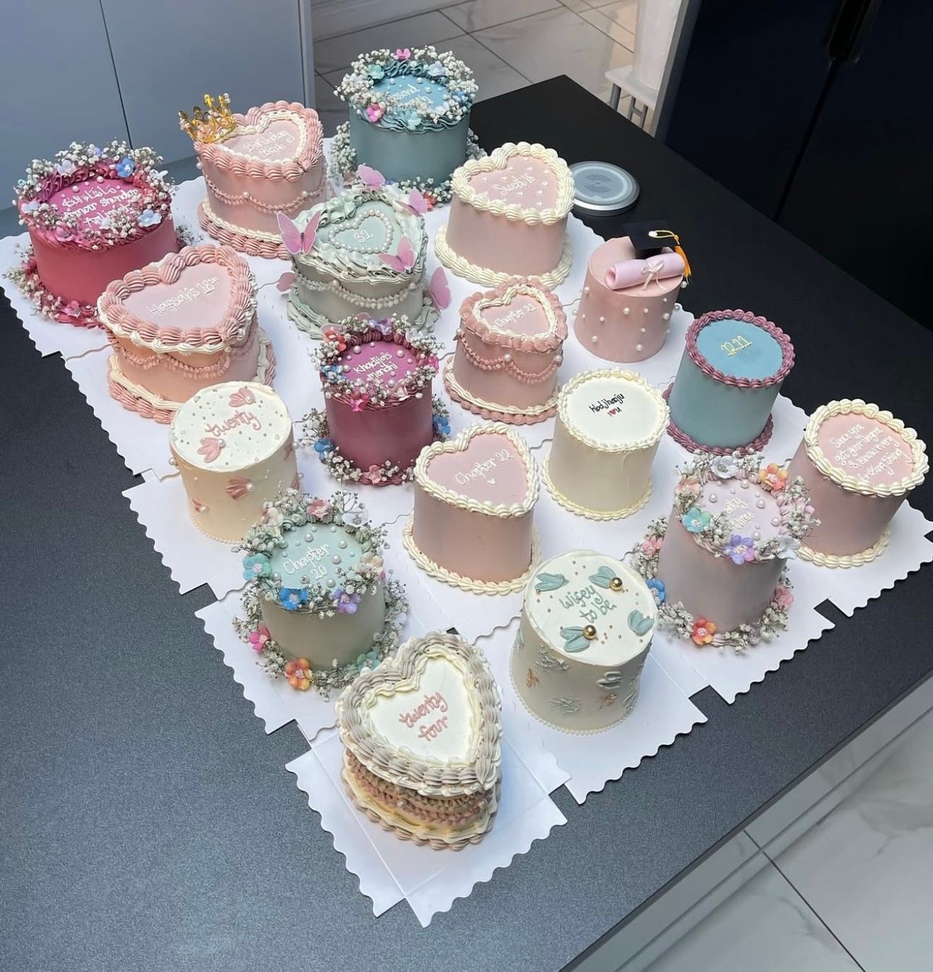 Assorted mini decorated cakes in pastel colors with floral and heart designs displayed on a white board.
