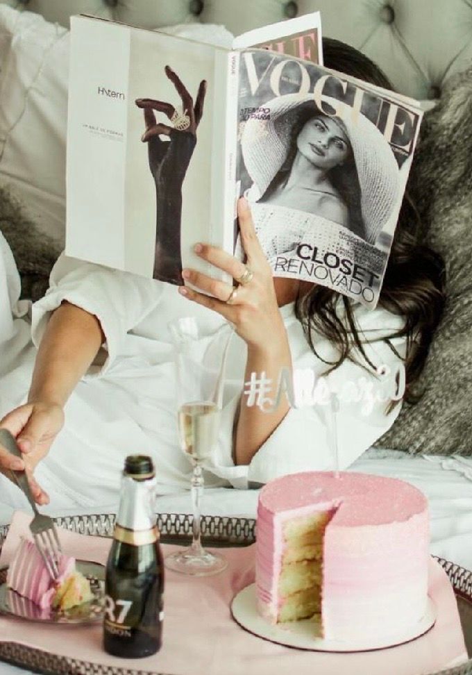 Woman in white robe reading Vogue magazine in bed with champagne and pink ombre cake.