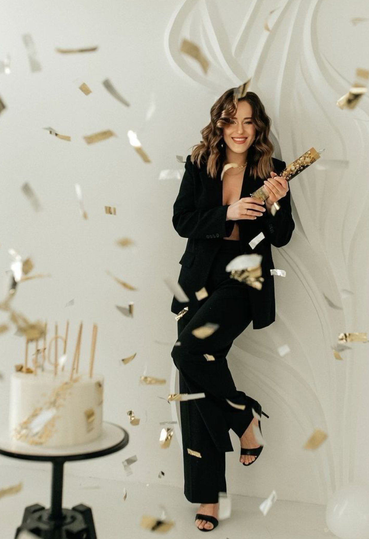 Woman in black suit celebrating birthday with confetti cannon beside elegant white cake with gold candles.