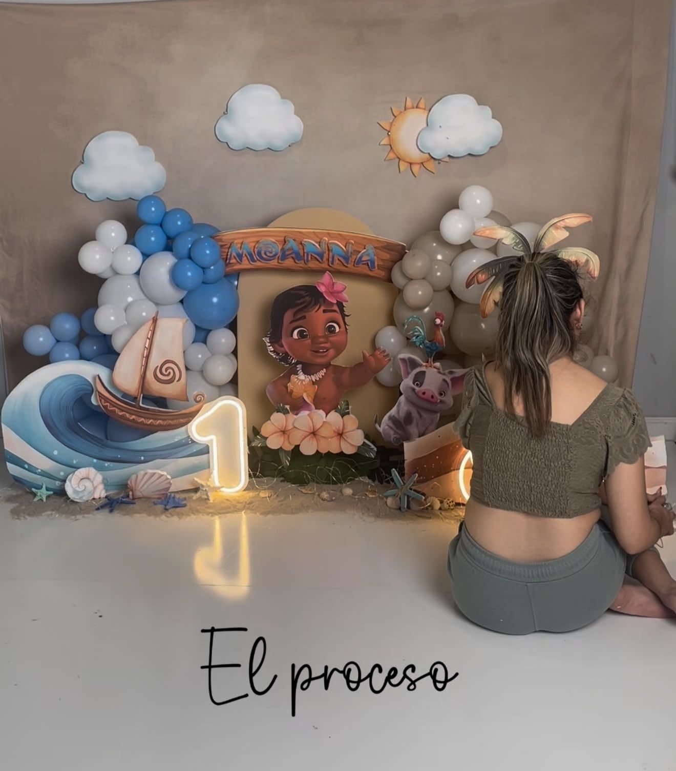 Girl sitting in front of a Moana-themed first birthday party backdrop with balloons and decorations.
