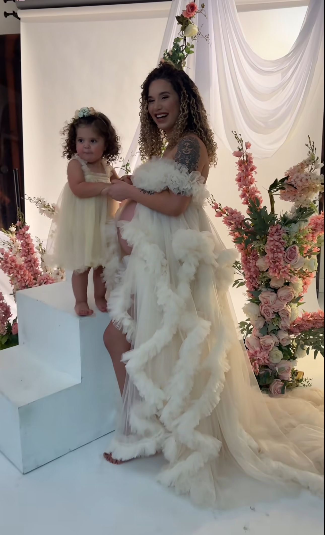 Woman in elegant white ruffled gown holding a toddler girl in ivory dress at a floral wedding ceremony backdrop.