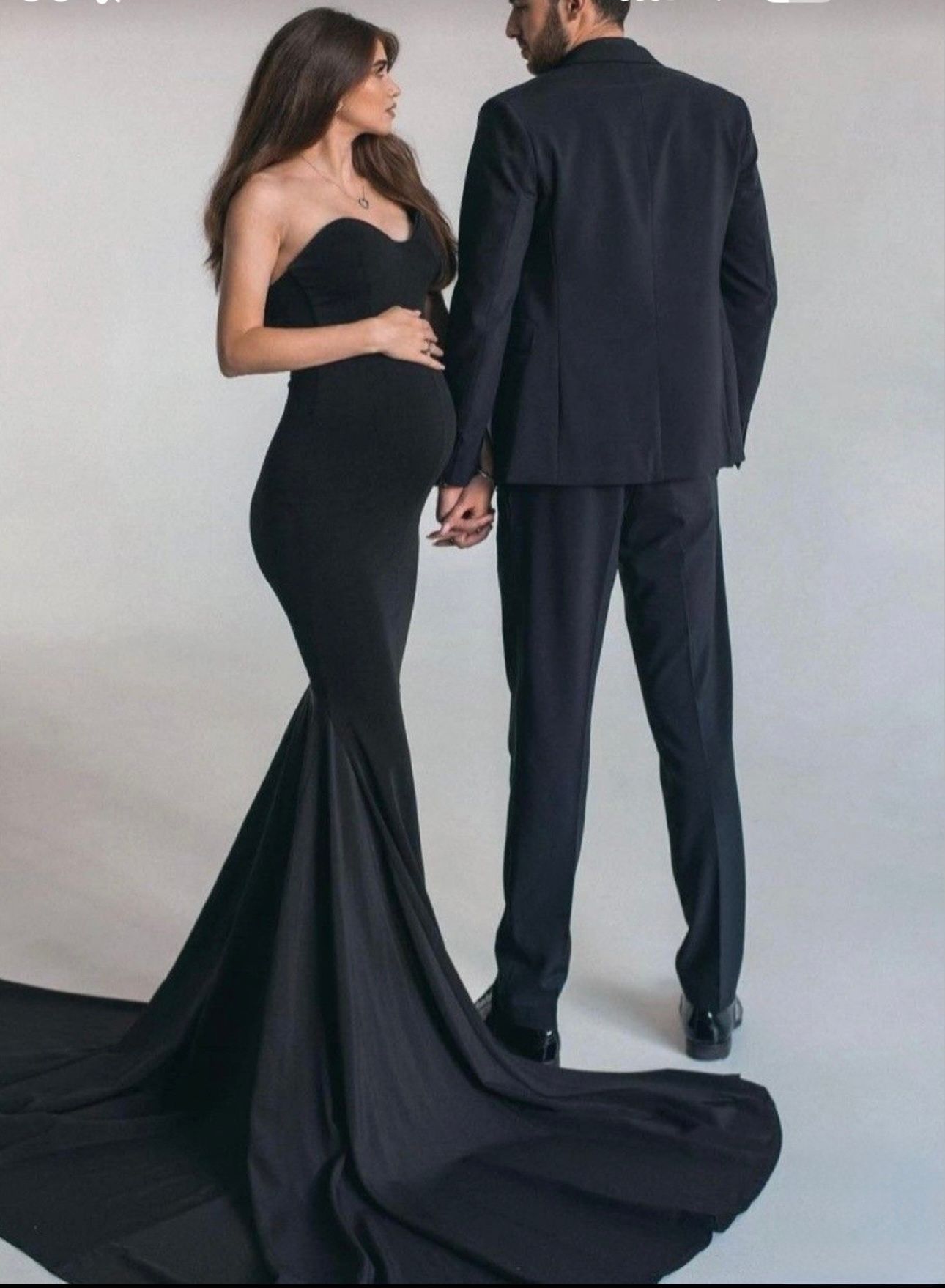 Pregnant woman in a black strapless mermaid gown holding hands with a man in a black suit in a studio.