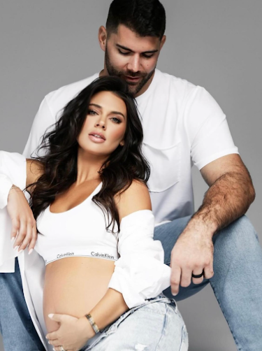 Intimate maternity portrait with couple in casual white and denim outfits against grey background.