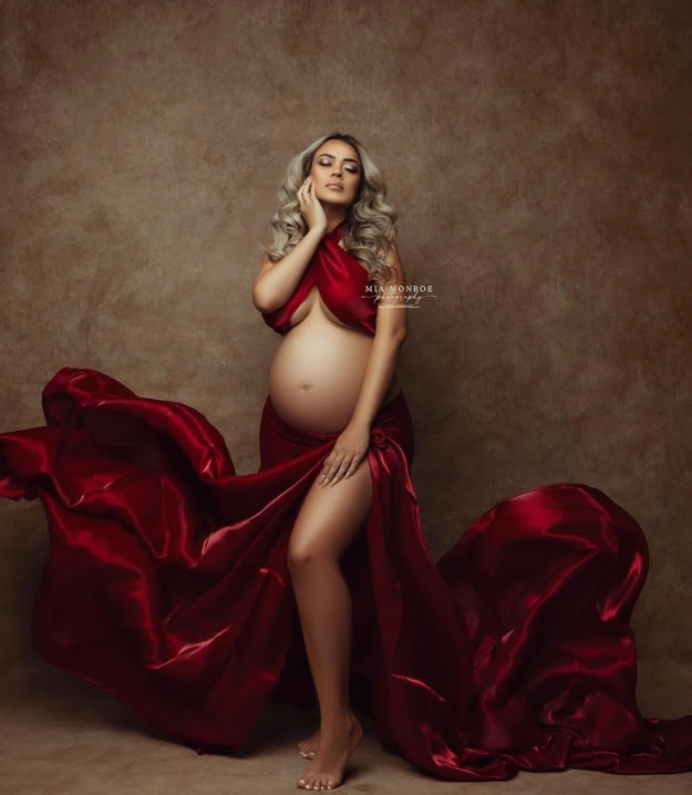 Elegant maternity portrait with flowing deep red silk dress against textured brown backdrop in dramatic studio lighting.