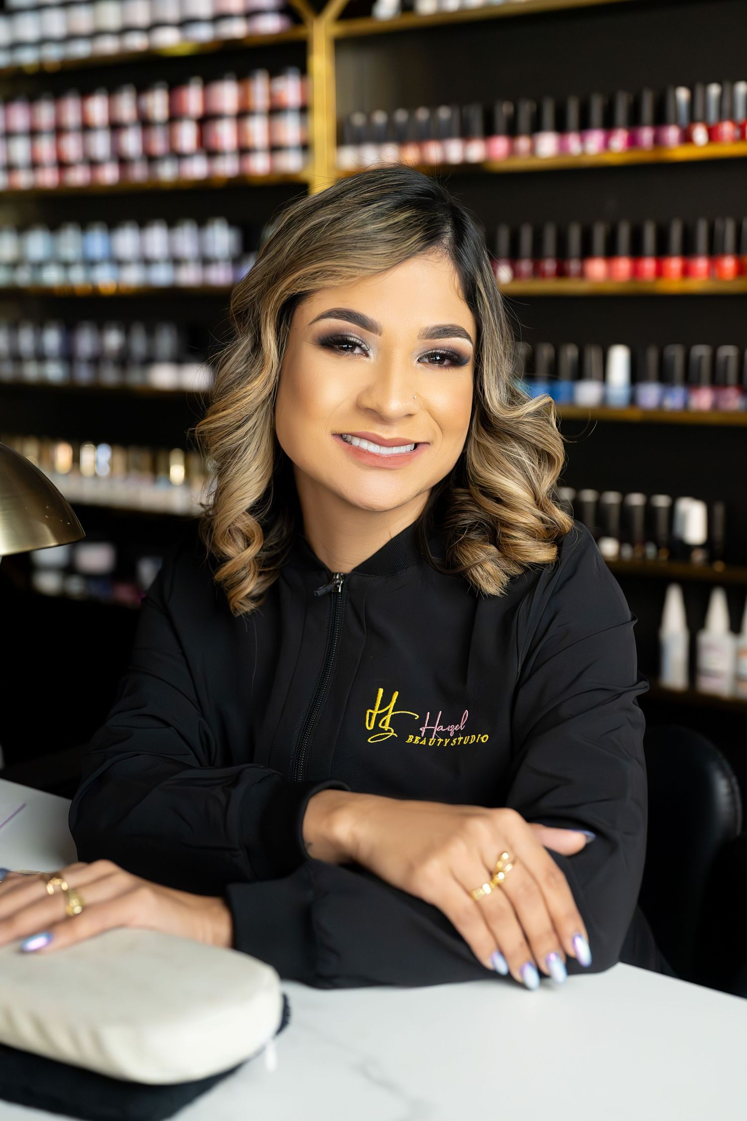 Professional in black uniform smiling at nail salon workstation with polish display wall behind.
