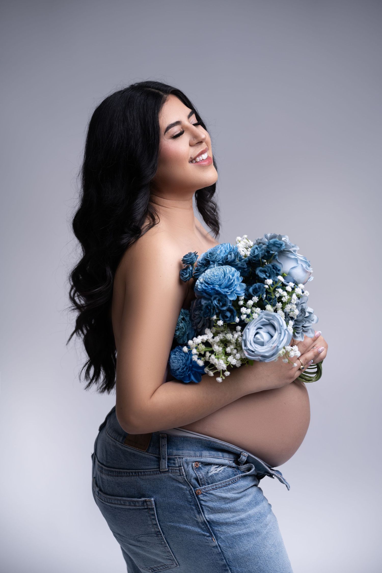 Maternity photo of an expectant mother holding blue roses and baby's breath bouquet against grey backdrop.