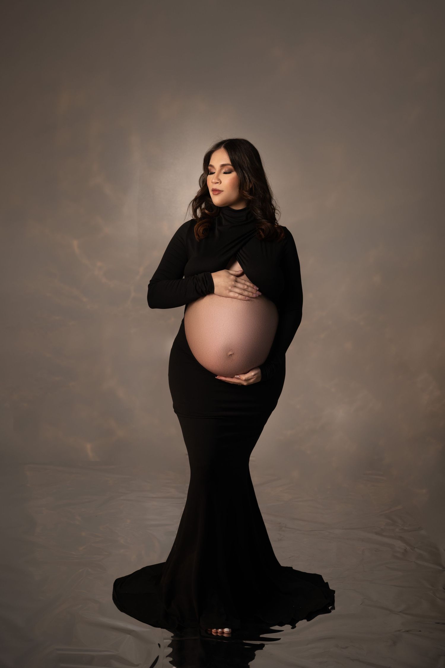 Moody maternity portrait in black fitted dress against dark atmospheric background.