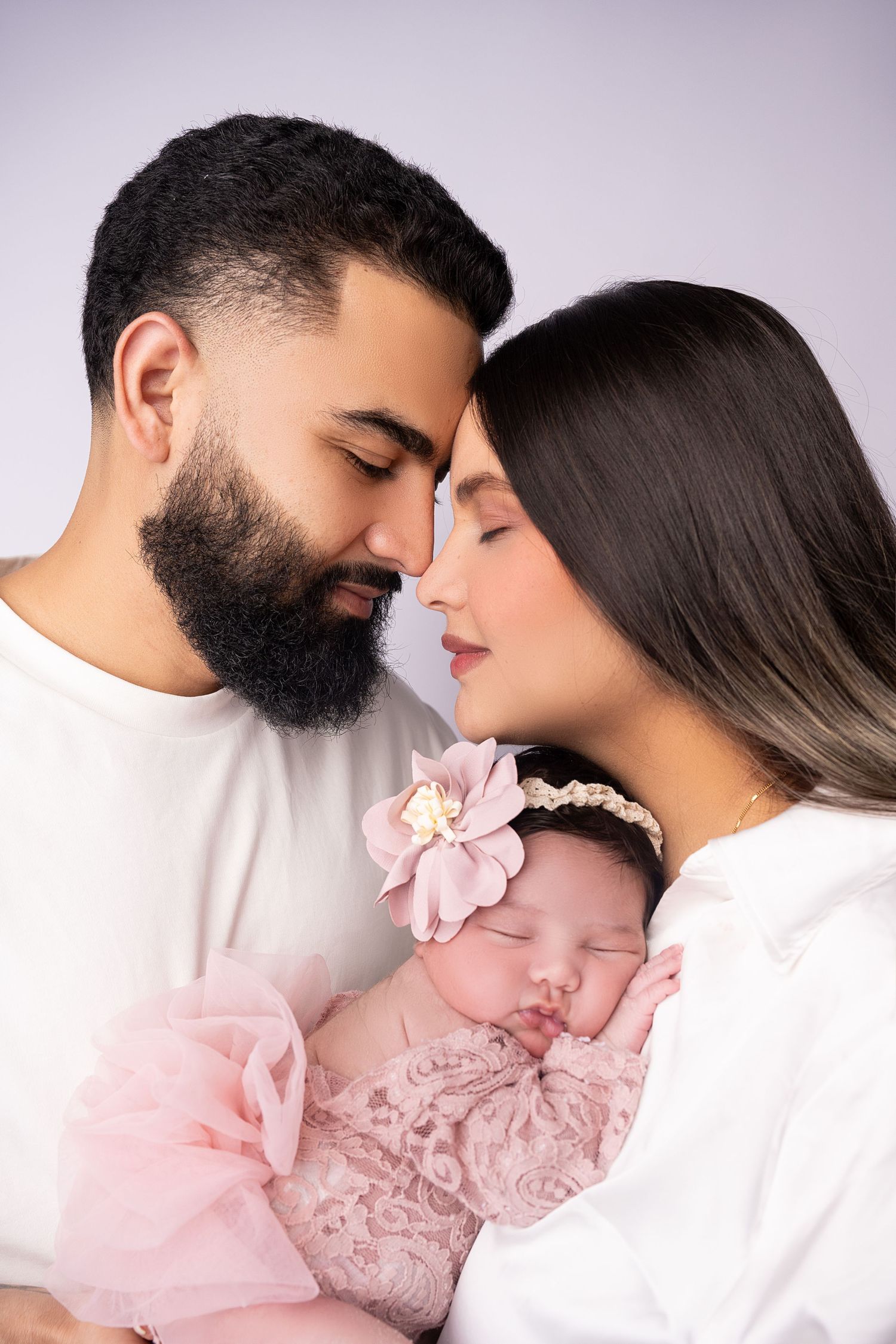 Intimate family moment with sleeping baby in pink lace against soft purple background.