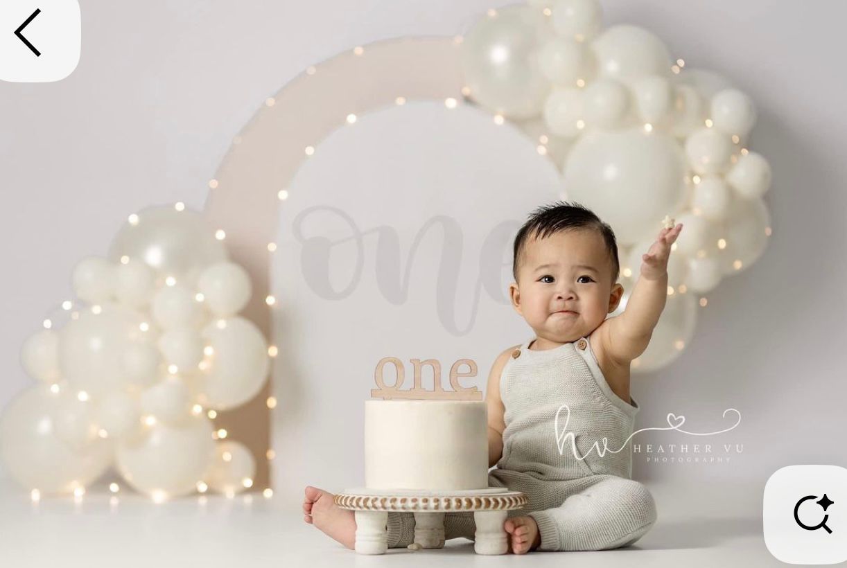 Baby's first birthday cake smash photo with white balloon arch, fairy lights, and “one” sign on neutral backdrop.