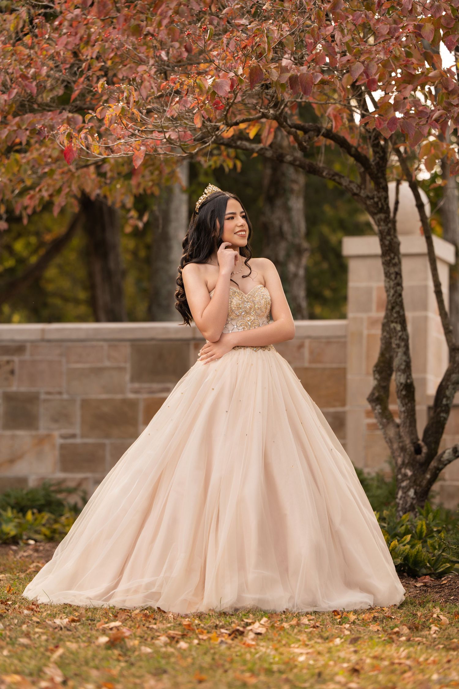 Young woman in champagne ball gown and tiara poses gracefully under autumn tree for quinceañera portrait.