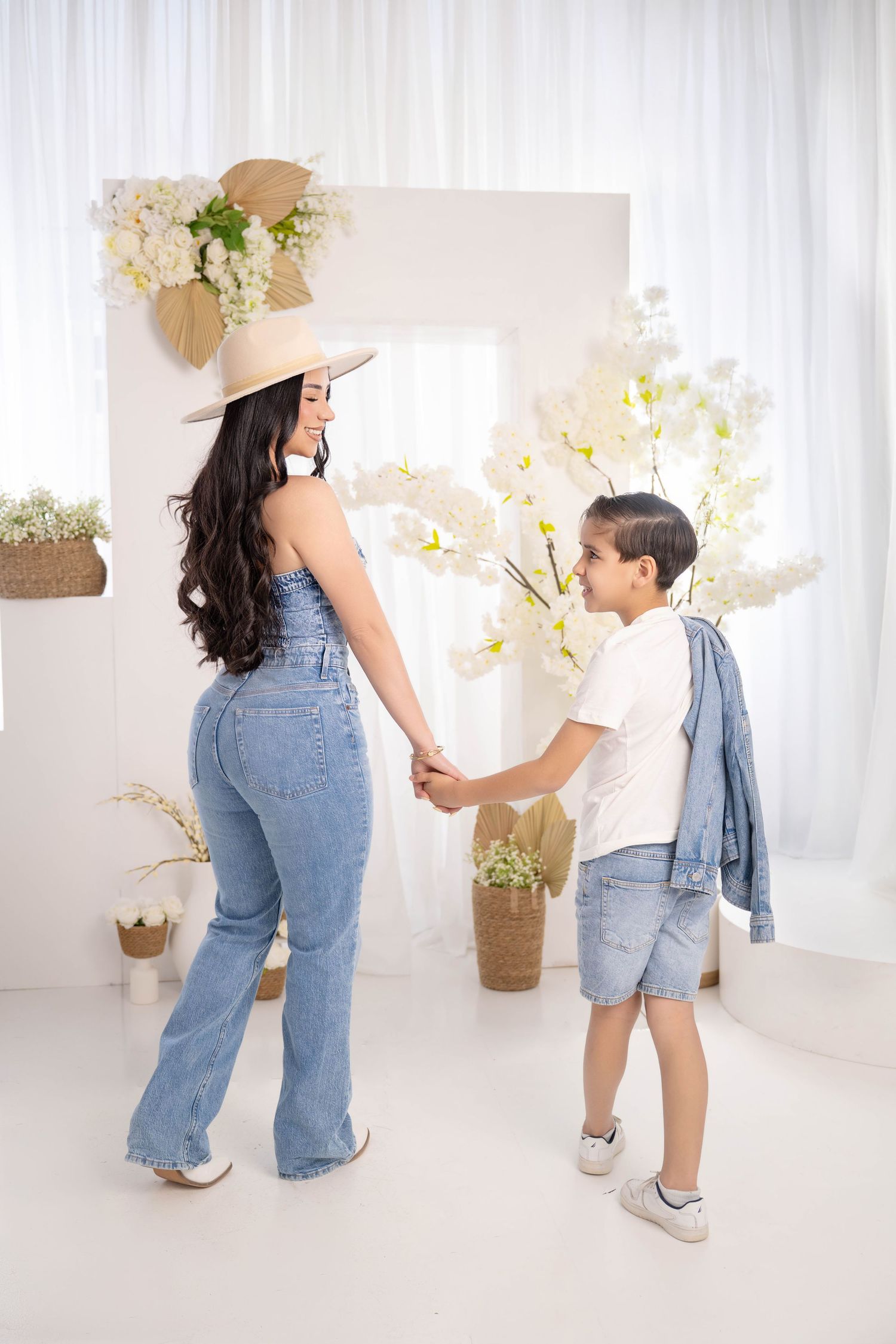 A woman in denim overalls and straw hat holds hands with a young boy in a bright white floral decorated room.