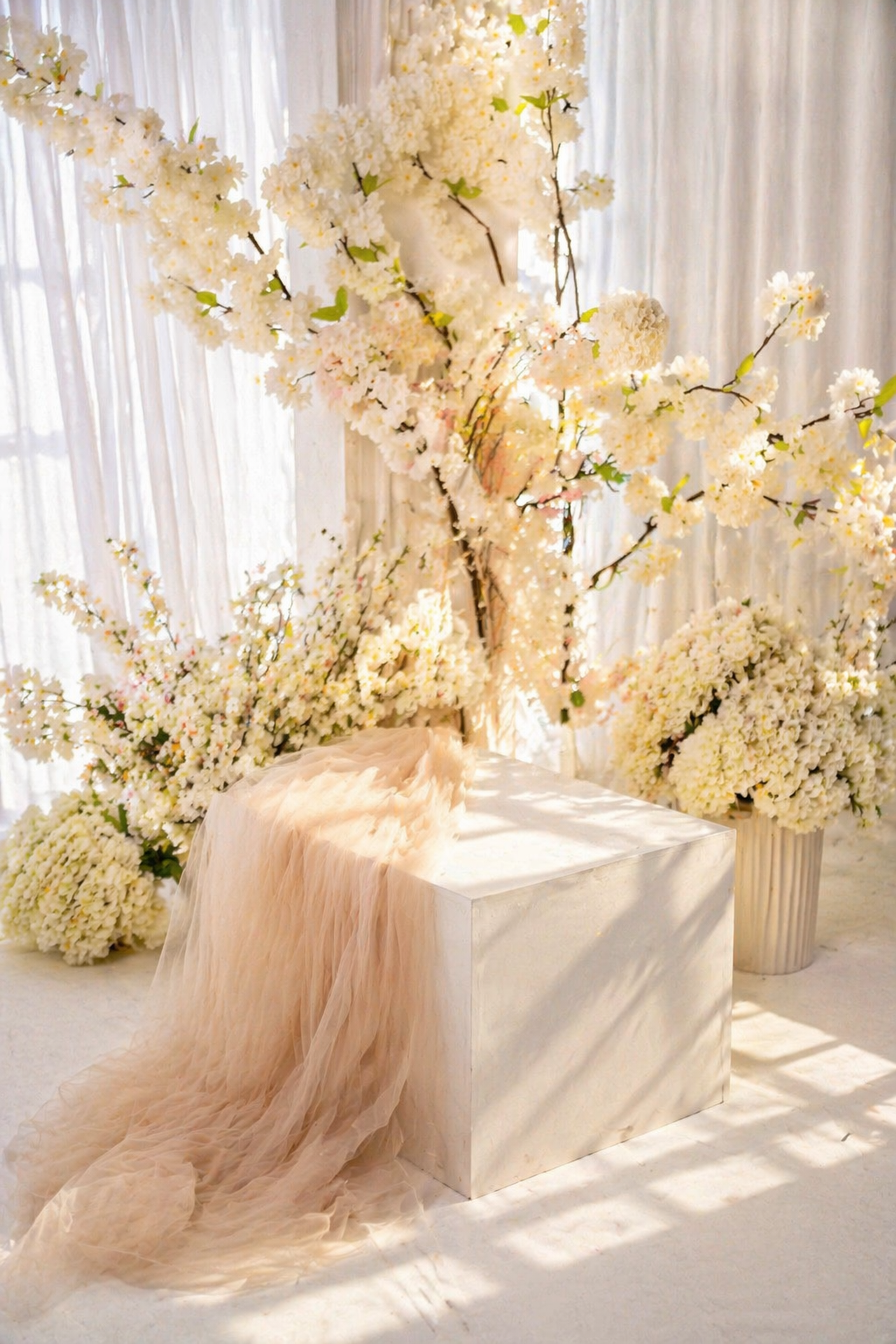 Elegant wedding decor with white cherry blossom trees, ivory florals, and a blush tulle drape over a white pedestal.