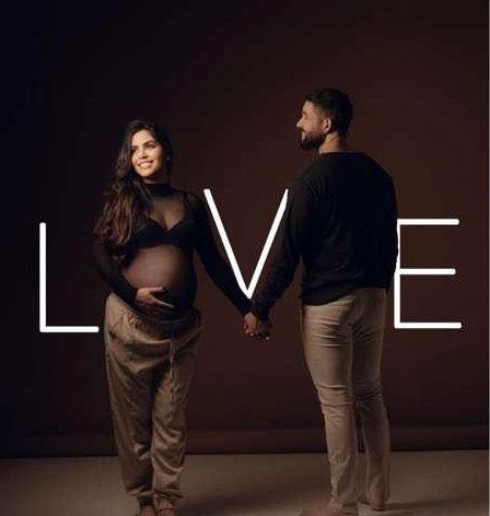 Pregnant couple holding hands with LOVE text overlay in a dark studio maternity photo shoot.