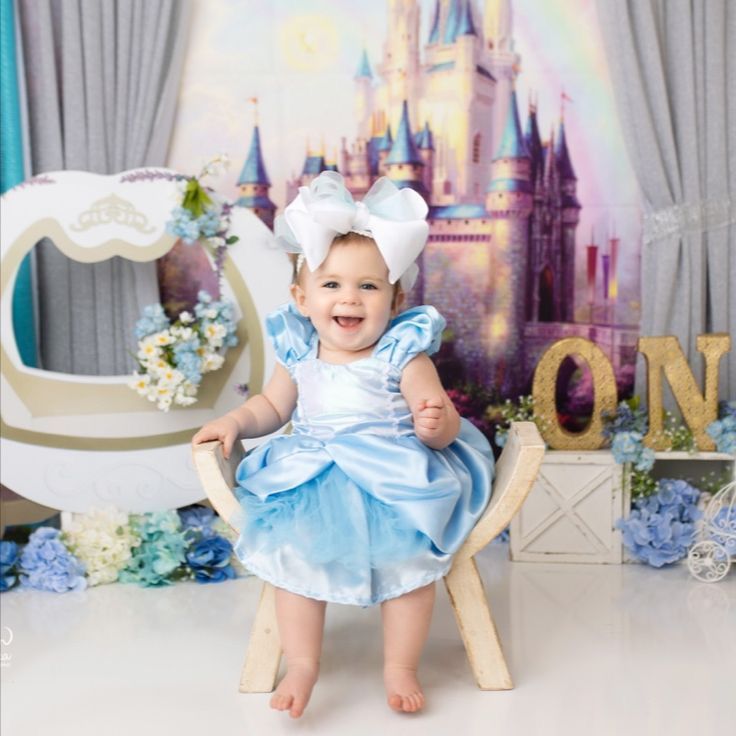 Baby girl in Cinderella blue dress with bow headband smiling in front of Disney castle backdrop.