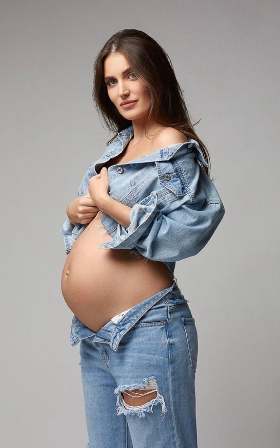 Pregnant woman in ripped jeans and denim jacket showing baby bump, posing against gray background.
