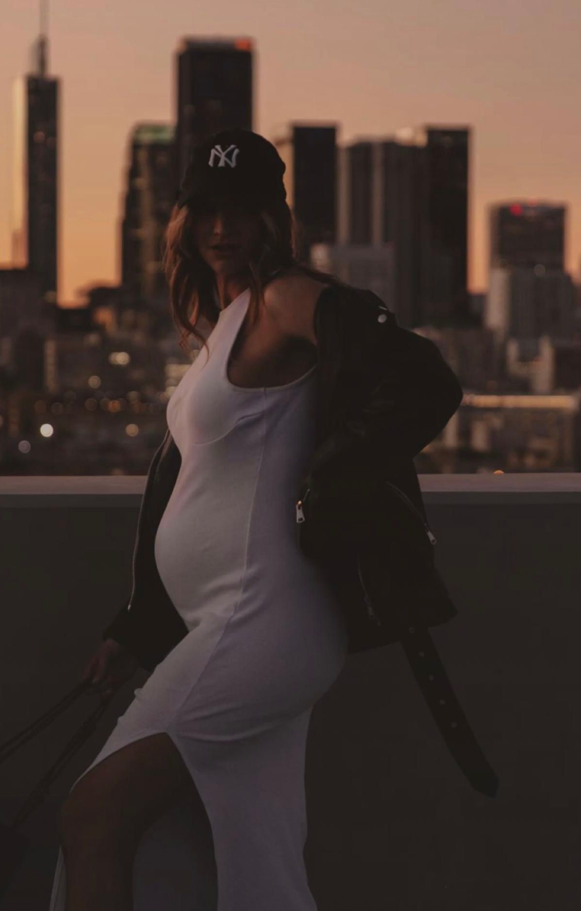 Pregnant woman in white dress and NY cap poses on rooftop at sunset with city skyline backdrop.