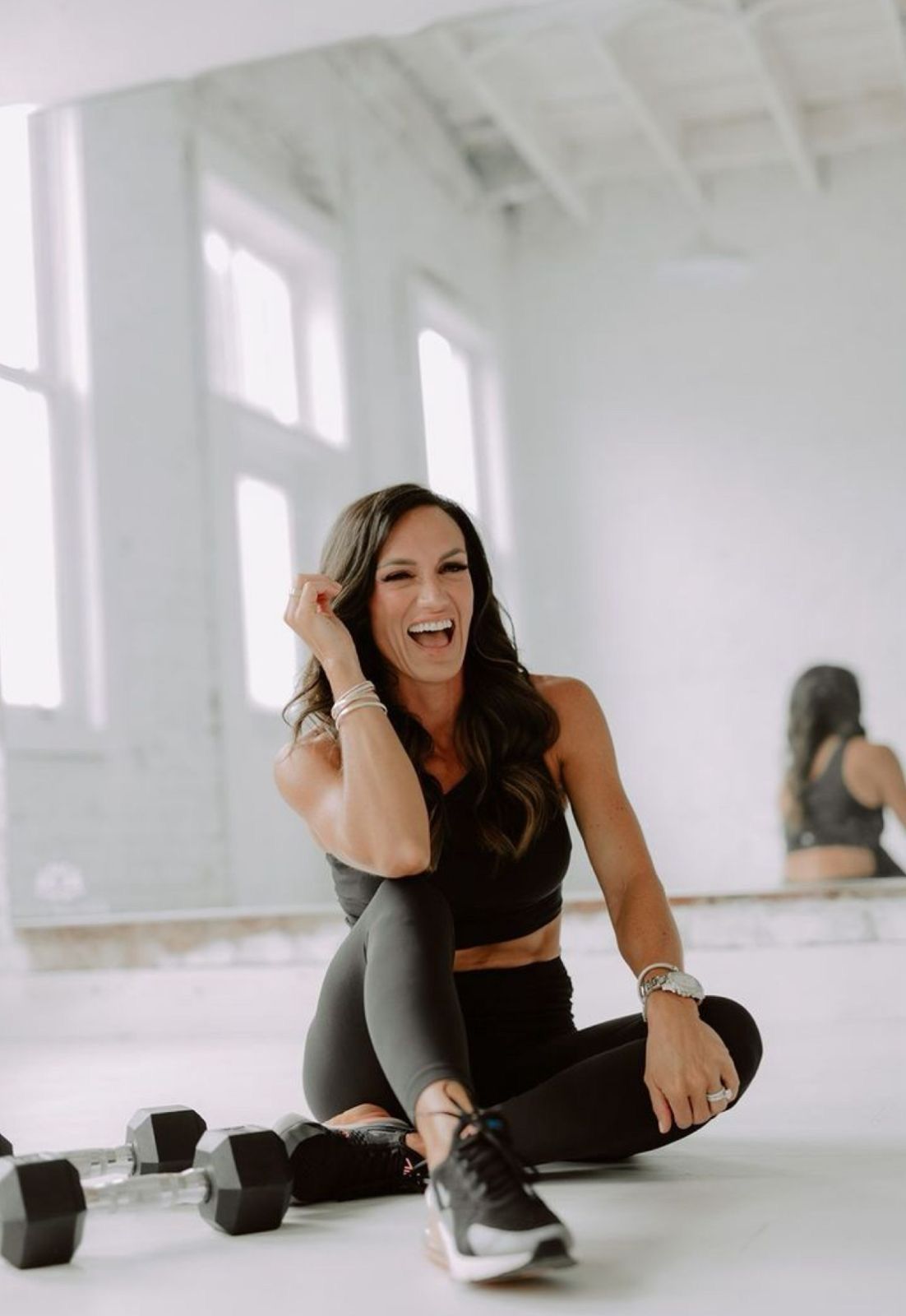 Fit woman in black workout set laughing while sitting on gym floor next to dumbbells.